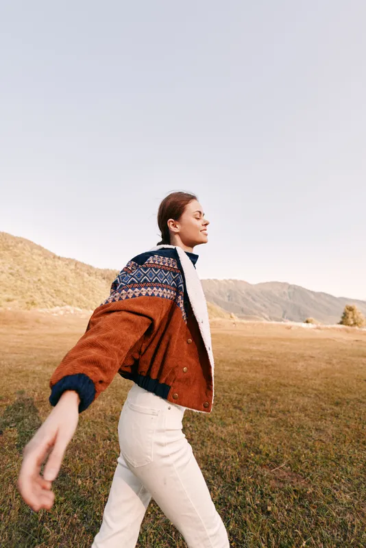 Woman walking in countryside field wearing patterned jacket and cozy sweater, enjoying autumn outdoors near mountains with sunlight, relaxed casual style and free travel lifestyle.