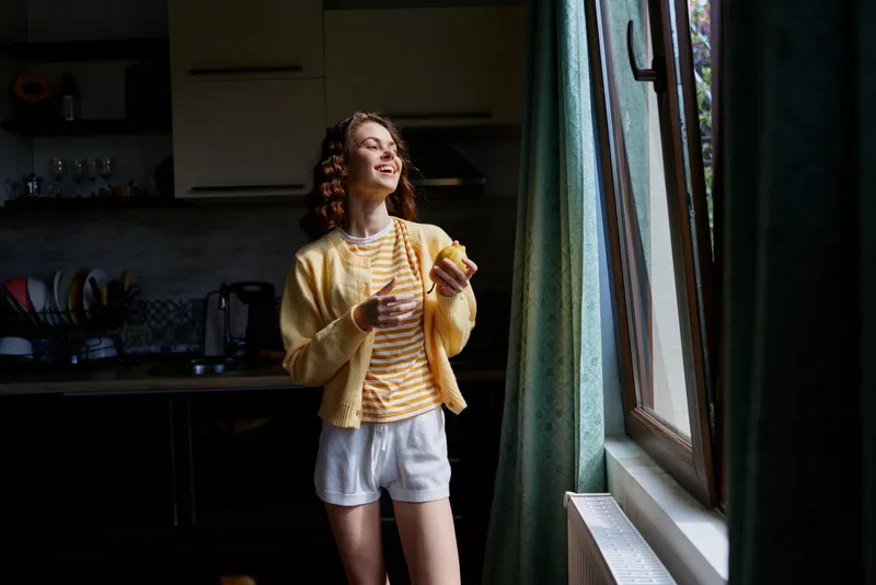 Bright young woman in a striped yellow cardigan stands in a sunlit kitchen by a window, smiling as she holds a fruit in a casual, relaxed morning moment.