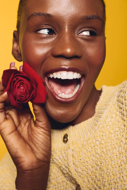 Smiling dark skinned African woman holds red rose near face, wearing yellow sweater. Close-up portrait on yellow background. Concept of beauty, happiness, and natural skin glow.