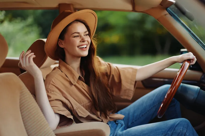 Happy young woman driving a vintage car, wearing a stylish beige outfit and a wide-brimmed hat, enjoying a sunny day outdoors.
