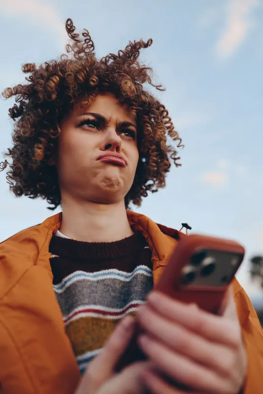 Young woman lifestyle portrait outdoors, curly hair in wind, holding smartphone near face, orange jacket and striped sweater, thoughtful gaze at camera, natural light on city street.