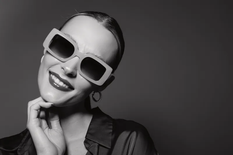 Smiling caucasian woman with fair skin wears large sunglasses and earrings, poses confidently on dark background. Concept of fashion, style, and beauty.