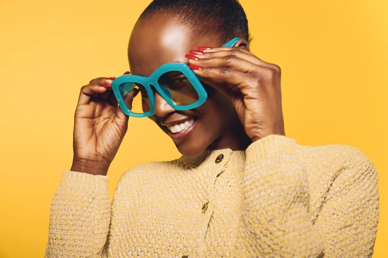 Happy African woman with dark skin wearing large blue sunglasses and beige sweater smiles against yellow background. Concept of fashion, style, and joyful lifestyle portrait.