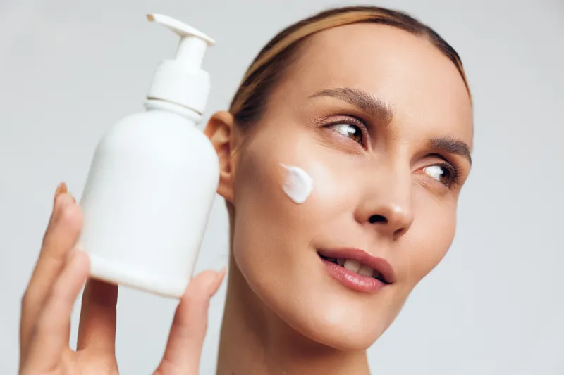 Young woman applying moisturizer cream on face holding white plastic bottle, skincare routine, clean fresh skin, natural beauty, soft light, neutral background.