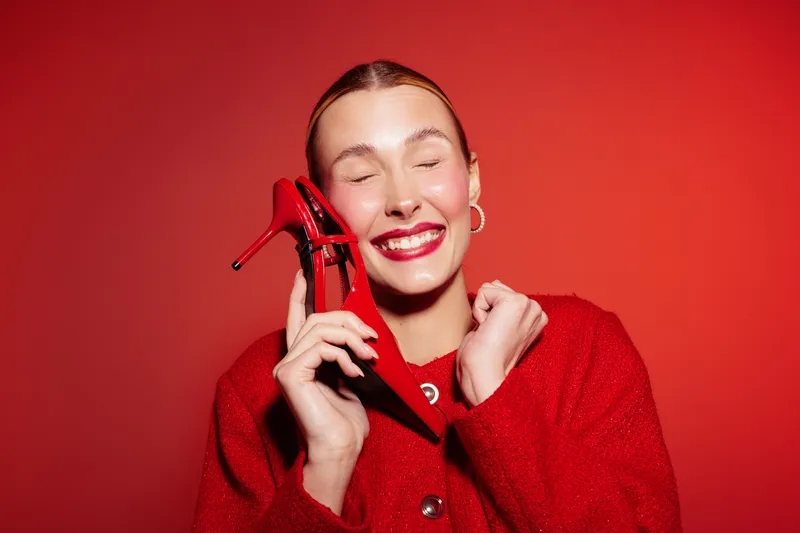 Happy woman in red sweater holds red high heel shoe near face with closed eyes and bright smile. Studio portrait on red background. Concept of fashion, joy, and style.