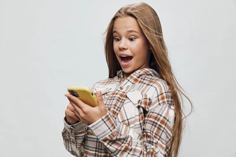 Girl with long hair and plaid shirt looking excited while holding yellow smartphone, surprised expression on face, isolated on light background with space for text.