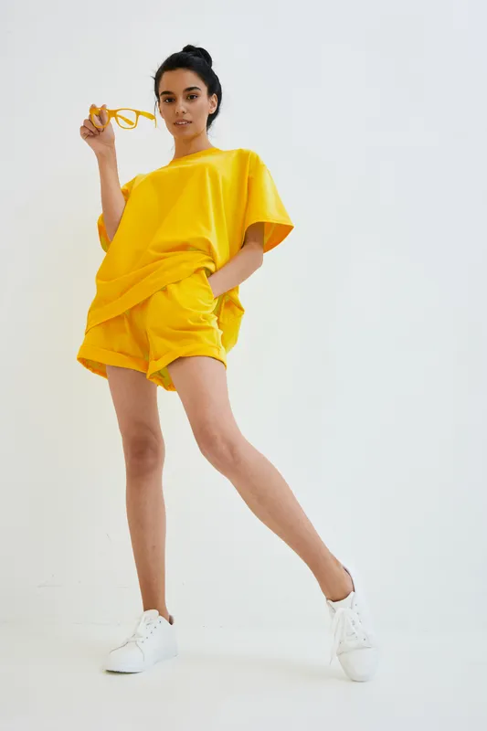 woman in yellow outfit standing confidently wearing shorts and t-shirt with white sneakers, holding sunglasses, modern casual style on white background