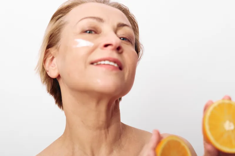 Mature woman with natural skin applies white facial cream, holding fresh orange slices. Bright studio lighting emphasizes skincare, healthy lifestyle, and vitamin c beauty concept.