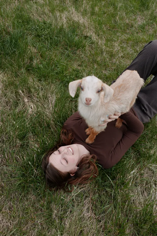 A young woman lies on grass smiling as she holds a small white baby goat on her chest in a relaxed outdoor lifestyle scene.