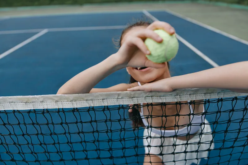 A joyful girl in a white sports outfit playfully holds a tennis ball, smiling through the net on a blue tennis court, capturing the fun of childhood and sports