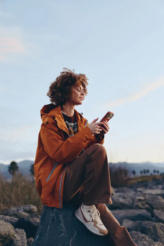 Woman lifestyle outdoors, curly haired woman sitting on rock, orange jacket using phone at sunset, relaxed pose and thoughtful smile, wide sky with soft clouds distant hills, evening breeze
