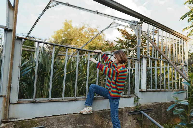 Eight-year-old girl child climbing a metal greenhouse fence in a suburban garden, wearing a colorful striped sweater and jeans, curious and focused expression during outdoor play.