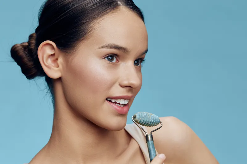 Young woman using a jade roller on her face, smiling and showcasing a fresh skincare routine against a blue background for beauty and wellness concepts