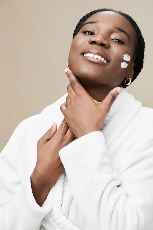 A young Black woman in a white robe smiles while applying skincare cream on her face, embodying a self care moment with a warm, neutral background