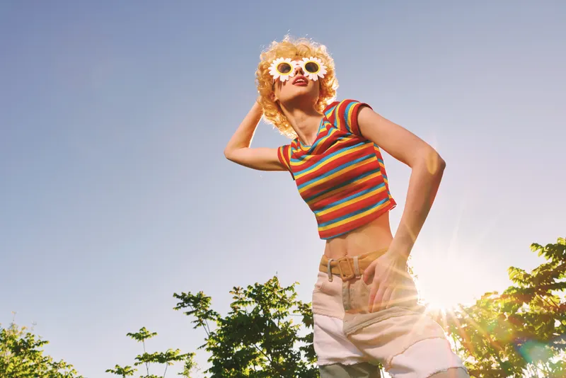 Young woman wearing a striped crop top and straw hat poses confidently outdoors under bright sun, enjoying warm weather, playful energy, and a carefree summer moment.