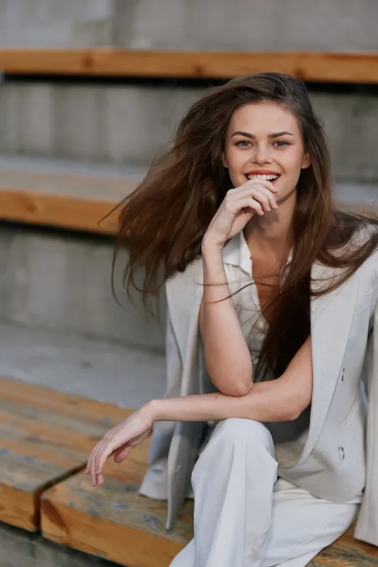 Confident young woman with brown hair in light beige business suit sitting on wooden steps smiling natural emotions
