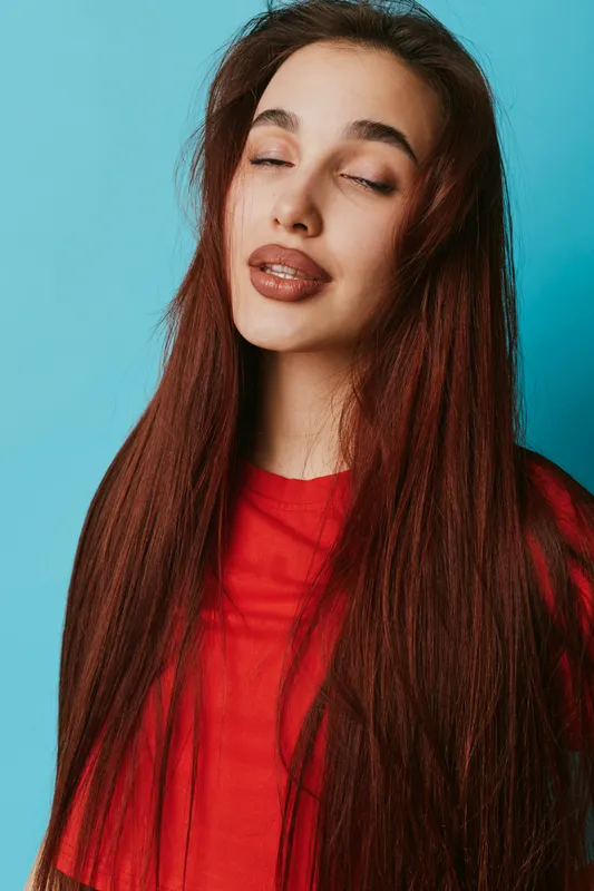 Young woman with long brown hair and freckles wearing red shirt posing with closed eyes and relaxed face against blue background in studio portrait. Beauty, fashion and casual style.