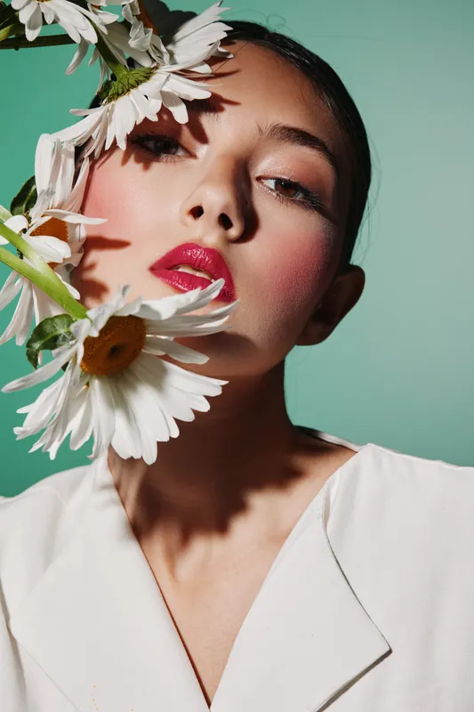 Fashion model with glossy pink lips posing with white daisies against a bright turquoise background. Close-up beauty shot focusing on makeup and natural flower elements. Creative concept.