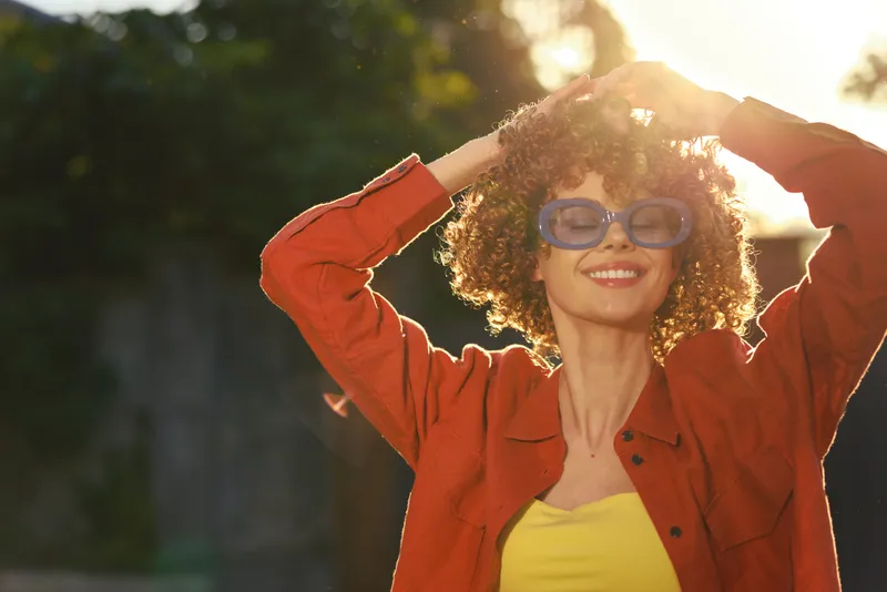 Smiling woman with curly hair wearing sunglasses and an orange jacket, outdoors in sunshine. A vibrant lifestyle portrait conveying warmth, confidence, and carefree energy