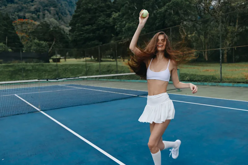 Cheerful young female tennis player in a white outfit enjoying a game on the court with a bright smile and energetic pose, surrounded by lush green nature