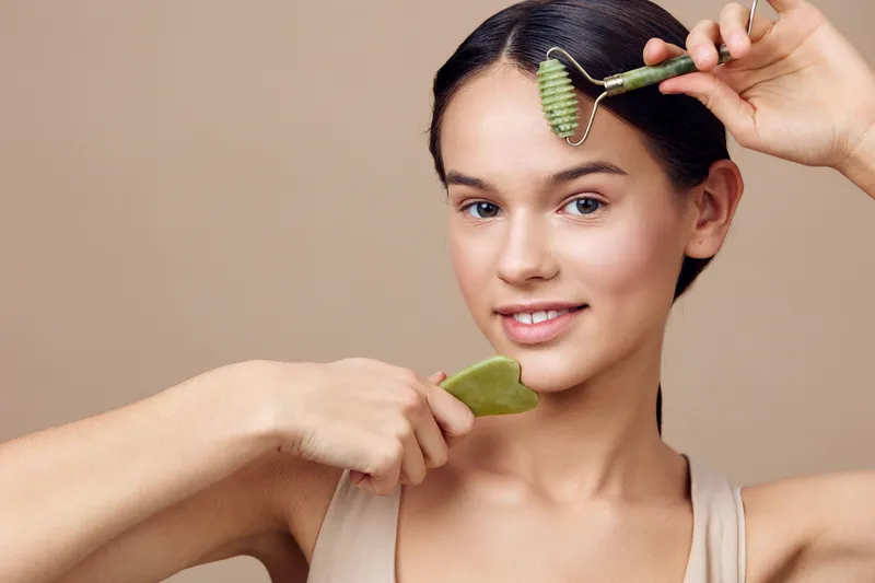 A young woman with a serene expression uses a jade roller and gua sha stone on her face, promoting healthy skincare routines and natural beauty