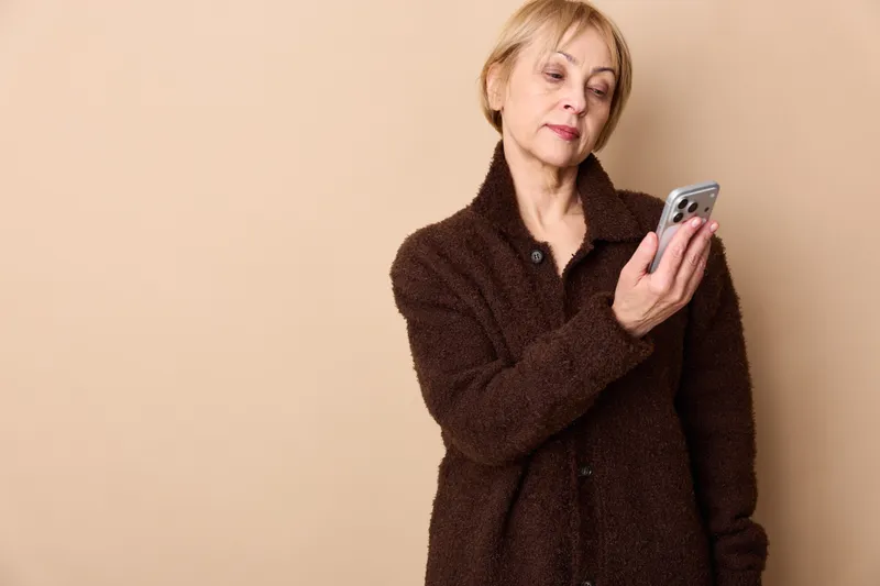 Woman senior holding smartphone for texting communication, technology portrait indoors in studio with beige background, calm mood, copy space for message today focus
