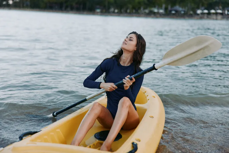 A young woman paddles a yellow kayak on calm water, wearing a navy swimsuit, with focused expression and graceful posture as she glides toward the shore.