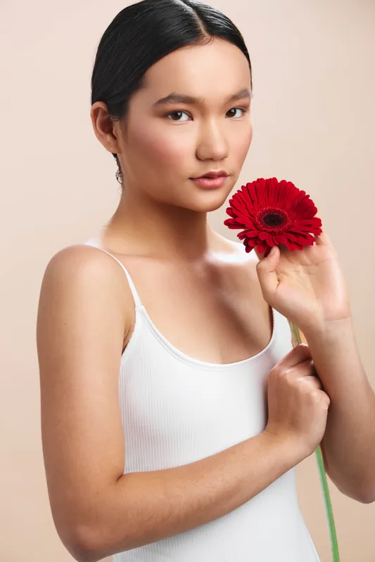 asian woman in a white tank holds a vivid red flower close to her cheek, smiling softly in a neutral studio setting, creating a warm, approachable portrait suitable for lifestyle and beauty themes.