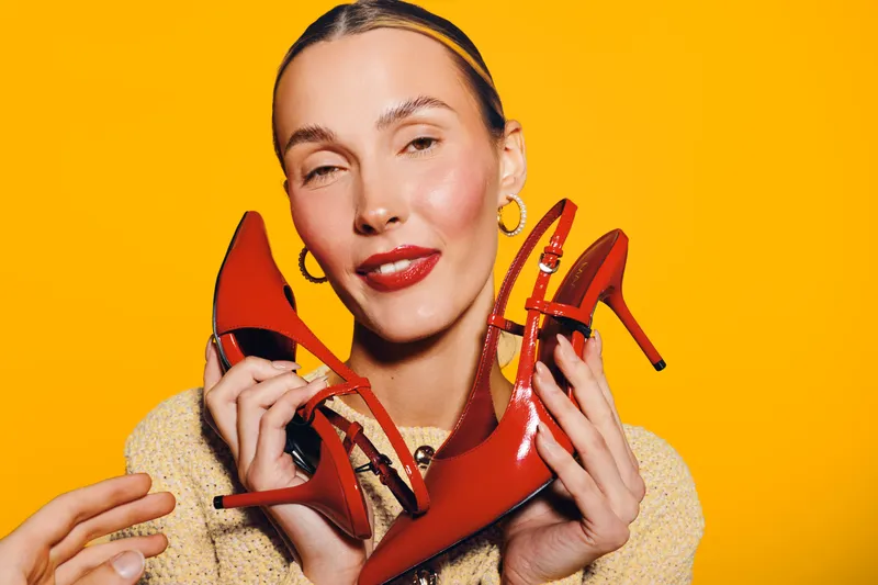 Young woman with slicked hair holds red high heel shoes in both hands on bright yellow background. Fashion concept with confident model wearing gold earrings and red lipstick.