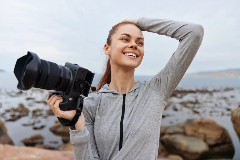Happy young woman holding a camera, smiling with a coastal background, wearing a casual grey outfit, showcasing a carefree and vibrant lifestyle