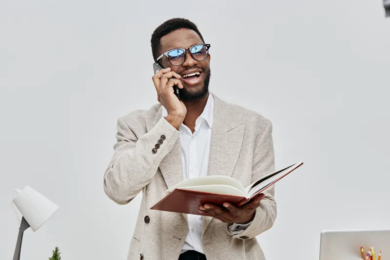 Smiling man wearing glasses talking on mobile phone hold open book in hand casual beige cardigan chic look studio portrait. People lifestyle concept