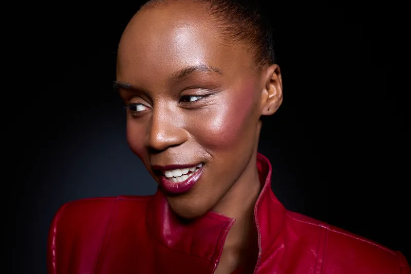 Close-up of a smiling dark skinned African woman wearing a red leather jacket looking sideways. Studio portrait with black background and shiny skin highlights. Concept of fashion and beauty.