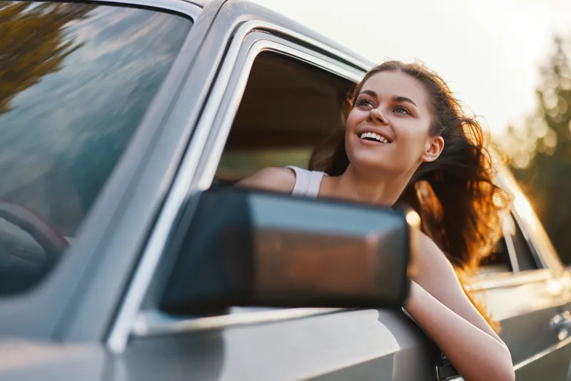 Joyful young woman looking out of car window on a sunny day, embracing freedom and adventure, with long hair flowing and a bright smile, perfect for travel themes.