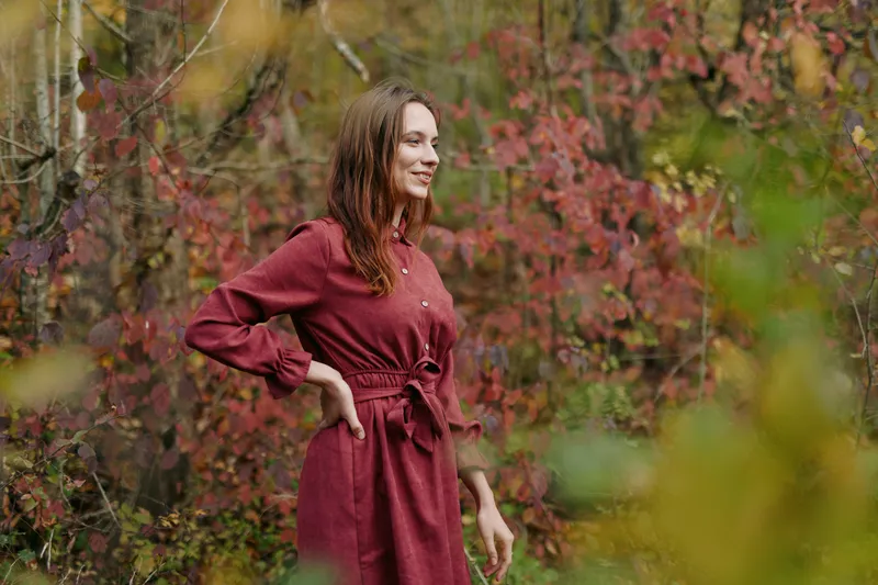 A woman in a burgundy dress stands in an autumn grove, radiating authenticity and credibility through relaxed posture, natural light, and a candid, genuine moment.