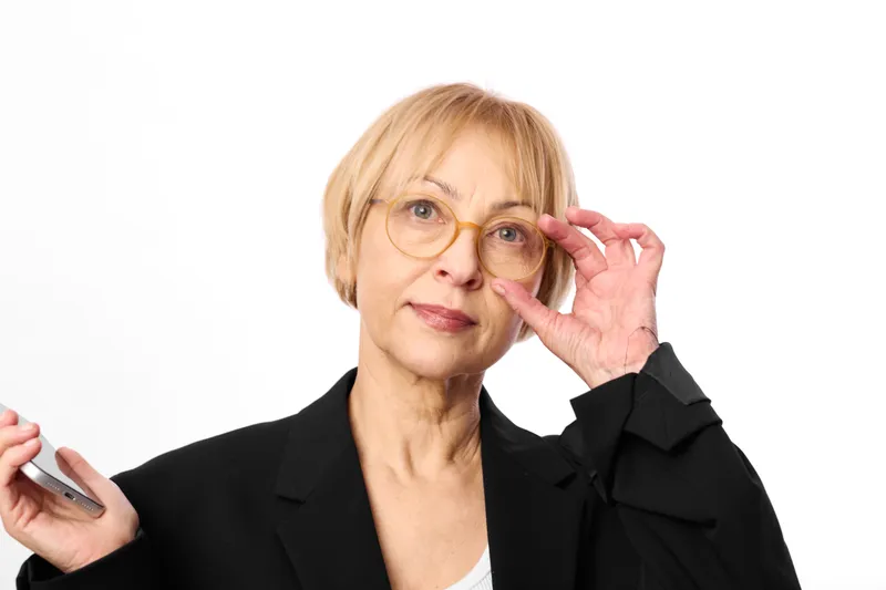 Elderly woman holding smartphone and glasses, adjusting frames with stressed expression. White background portrait, conveying anxiety and concern about communication.