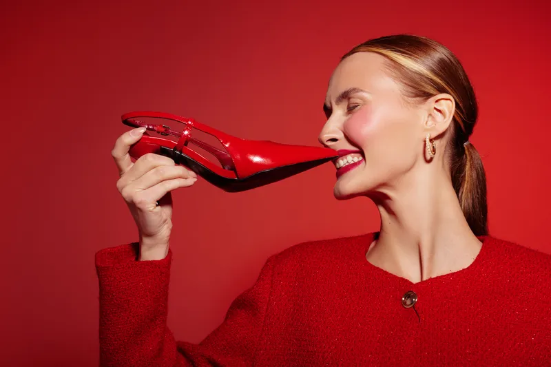 A smiling woman in red jacket holds a red high heel shoe close to her face against a red background. Concept of fashion, style, and bold color contrast in studio lighting.