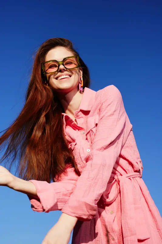 Cheerful young woman in a pink dress enjoying summer outdoors, against a bright blue sky, showcasing vibrant colors and joyful energy, perfect for fashion and lifestyle concepts