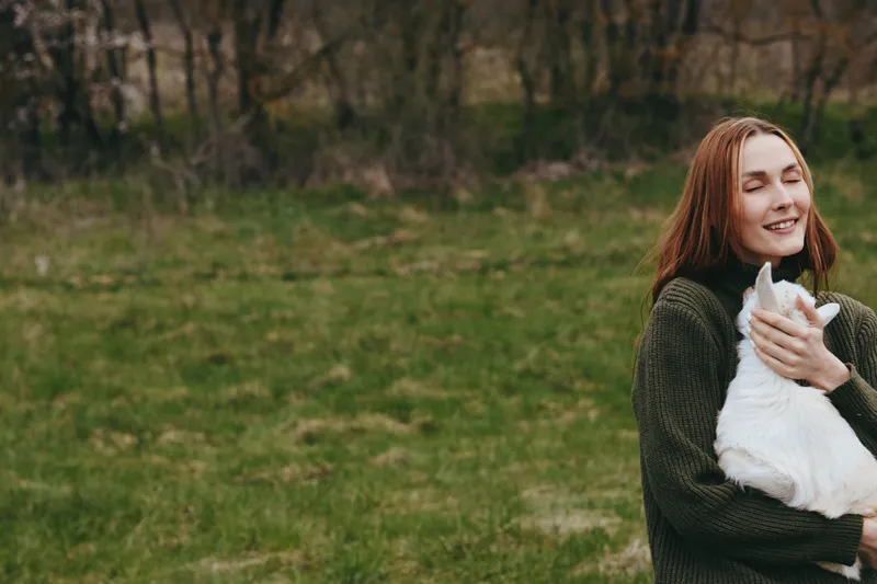 Young woman with long red hair smiles warmly while holding a white baby goat outdoors in a green grassy field during a relaxed lifestyle moment.