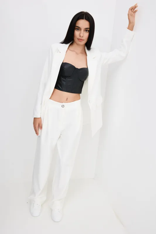 stylish woman in white pantsuit and black corset top posing indoors against white wall, modern fashion, minimalistic design, confident mood