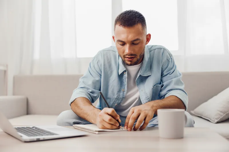 Focused man writing in a notebook, seated on a cozy couch with a laptop and coffee. A bright, airy environment enhances creativity and productivity.