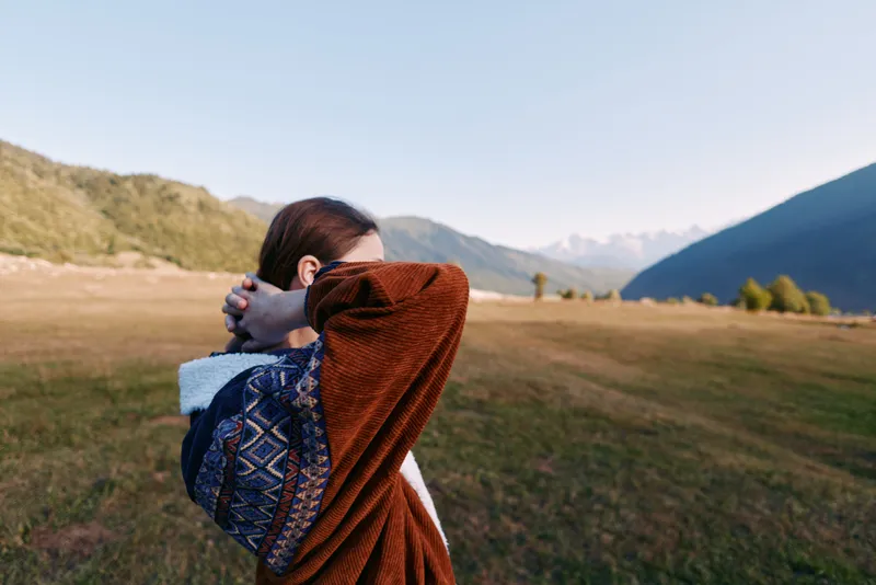 Woman in patterned sweater stretches and relaxes in nature on a grassy meadow with mountain landscape in the background, outdoors travel portrait capturing calm and freedom.