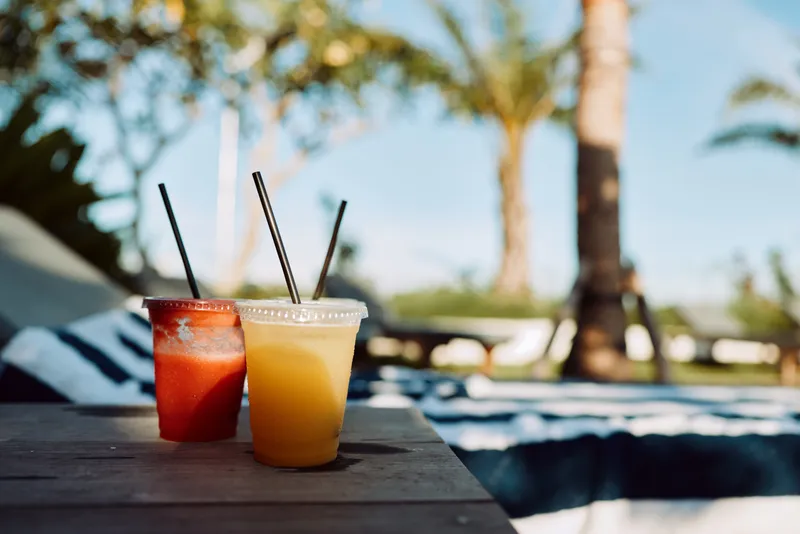 Two cold beverages in plastic cups with straws on a wooden table outdoors. Sunny day with blurred palm trees and blue sky in the background. Concept of summer refreshment and relaxation.