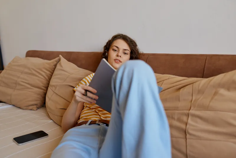 A young woman sits on a sofa at home, relaxing while reading a tablet and enjoying a calm casual moment in a cozy living room.