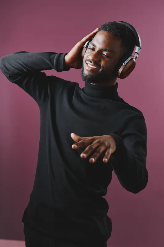 Young Black man enjoying music with headphones, smiling joyfully in a stylish black turtleneck against a vibrant purple background, conveying happiness and relaxation