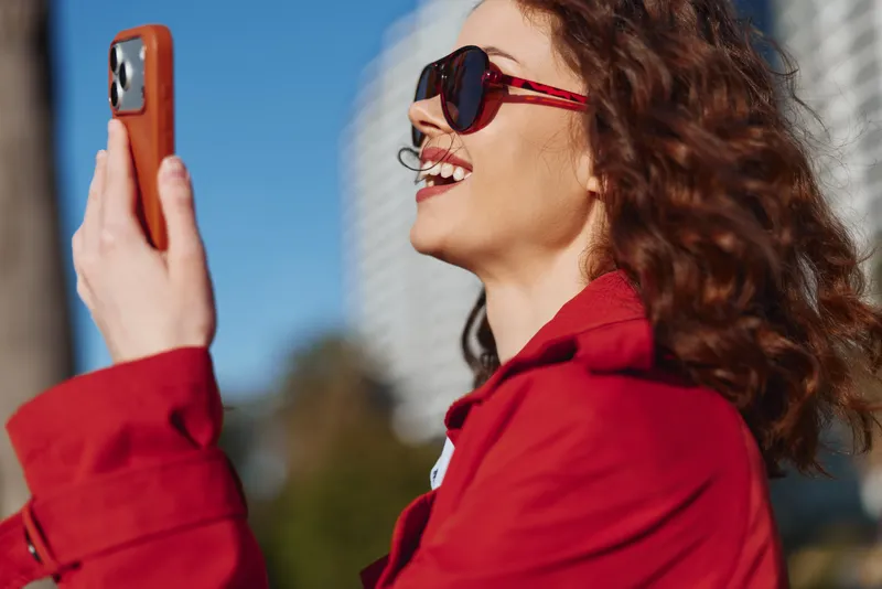 Smiling woman in a red jacket takes a selfie outdoors on a sunny day. Bright urban backdrop with blue sky and modern buildings. Fashionable, confident, lifestyle moment.