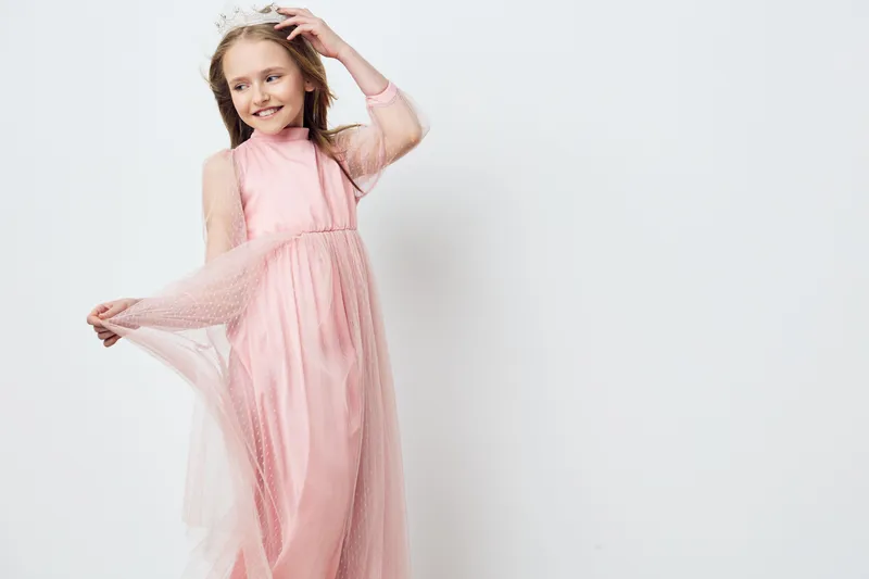 Cheerful young girl in a pink dress with a flowing skirt, dancing and playing joyfully against a light neutral background, evoking a sense of innocence and happiness