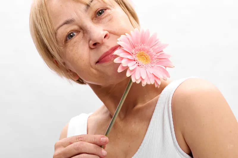 Portrait of a woman holding a pink gerbera flower near her lips, blond hair, soft light in studio, beauty and spring nature mood, calm smile, gentle femininity, concept