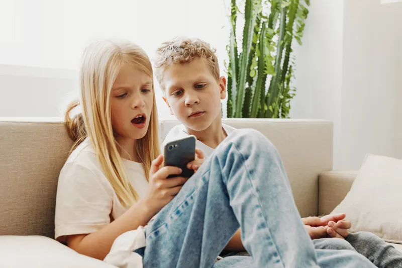 Curious children sitting on couch looking at smartphone screen together in bright living room. Concept of technology use, childhood curiosity, and shared digital experience in home environment.