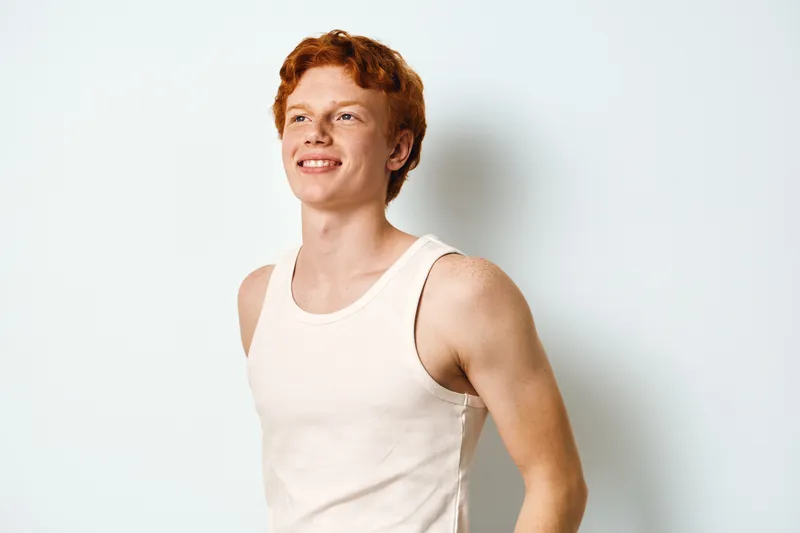 Young man with red hair smiling and wearing white tank top standing against white background looking away. Portrait of happy male with casual clothes and relaxed pose indoors.
