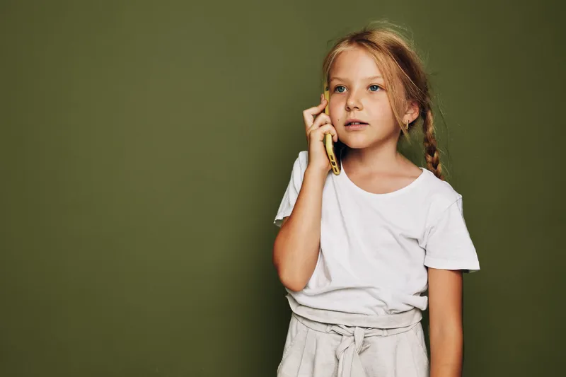 Young girl with braided hair wearing white shirt talks on phone against green background. Casual child portrait with natural expression and simple clothing.